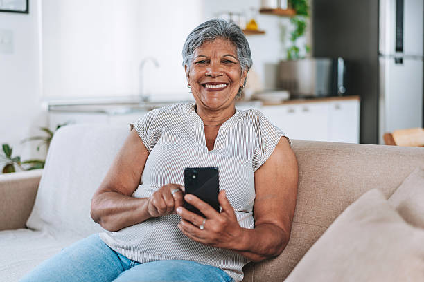 istock Smiling Senior Woman Sitting On Sofa Using Smartphone Looking at camera 2252563225