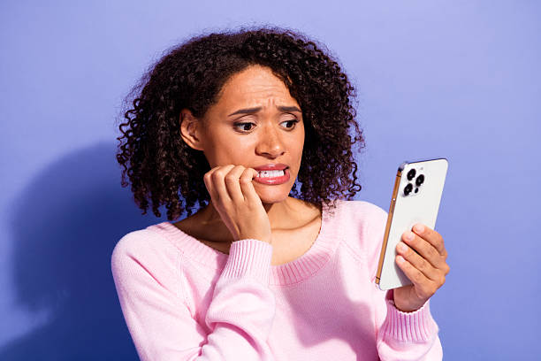 istock Young woman in pink sweater bites lip while looking at smartphone with worried expression against violet background 2252566982