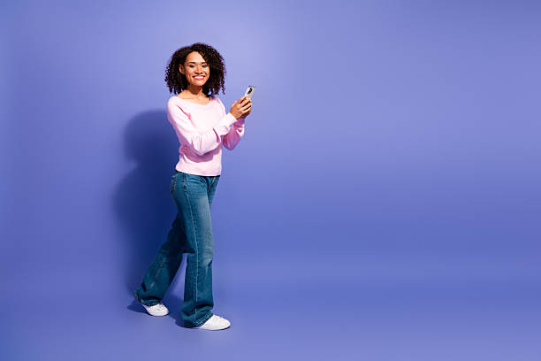 istock Young woman in pink sweater smiles while using a smartphone against a violet studio backdrop 2252567107