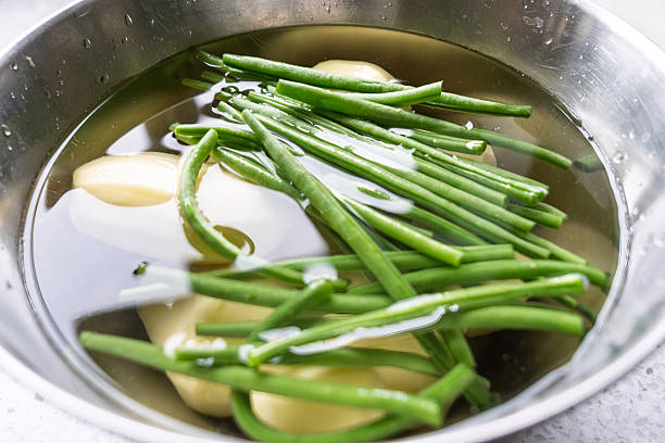 Close-up view of fresh green beans (Phaseolus vulgaris) and peeled potatoes (Solanum tuberosum) submerged in water inside a shiny stainless steel pot. The vibrant green pods and white potato chunks are partially visible through the water, with bubbles on the surface. This indoor kitchen scene captures the preparation stage of a simple vegetable dish, emphasizing freshness and simplicity. Ideal for commercial uses in cookbooks, recipe websites, healthy eating blogs, and food packaging to illustrate blanching techniques, vegetarian meals, or farm-to-table concepts. The image highlights natural ingredients without any people, suitable for editorial content on nutrition, seasonal produce, or home cooking tutorials.