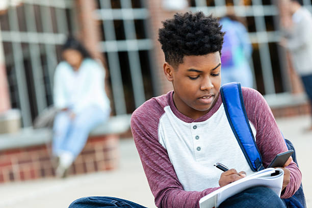 istock Young student focused on notebook, holding phone and pen outside school building. 2252574067
