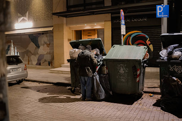 A person collecting items from garbage containers on a city street at night in Thessaloniki, Greece. The image documents urban poverty, social issues, and everyday life in a European city.
