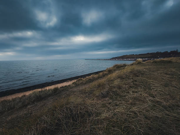 A wide-angle landscape of a moody, overcast sky with sunbeams breaking through dark clouds over the Oresund sea. The foreground features the sandy shore of Raa vallar beach covered in winter seagrass, with a long wooden swimming pier and lifebuoy station extending into the cold water.