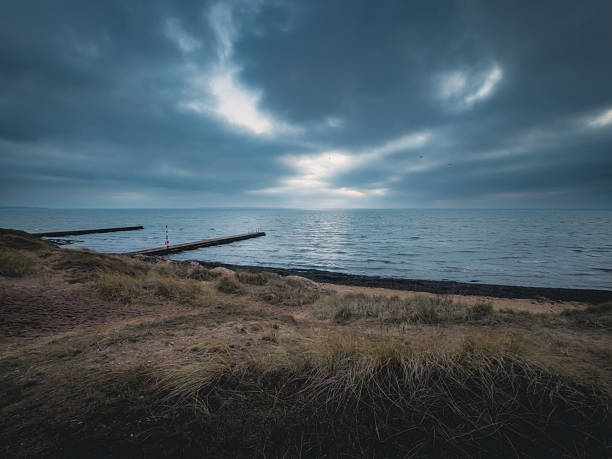 A wide-angle landscape of a moody, overcast sky with sunbeams breaking through dark clouds over the Oresund sea. The foreground features the sandy shore of Raa vallar beach covered in winter seagrass, with a long wooden swimming pier and lifebuoy station extending into the cold water.