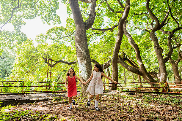 Sisters in colorful dresses run and play together in a vibrant park setting, surrounded by green trees and a wooden fence. Their movement and expressions capture the pure happiness and carefree spirit of childhood.