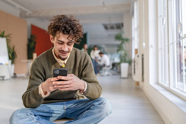istock Man sits on floor in modern office using smartphone while colleagues work nearby during business hours 2252595044