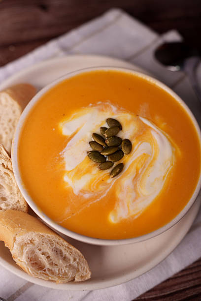creamy pumpkin soup garnished with green seeds and served with sliced rustic bread on a light ceramic plate, set against a wooden table background with soft lighting for a cozy ambiance, photography