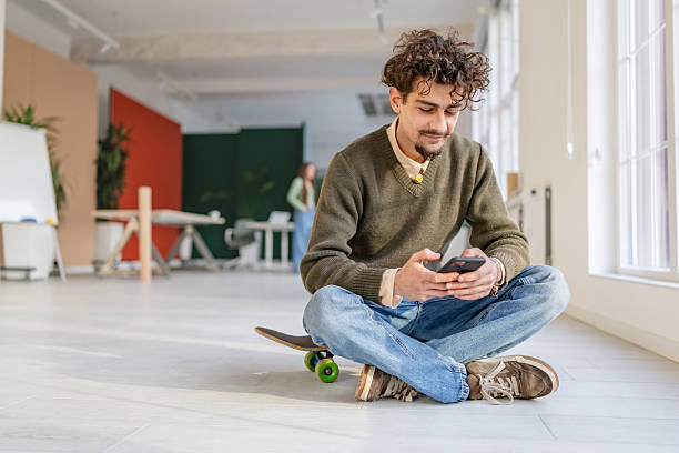 istock Young adult sits on skateboard in office space while using smartphone and enjoying casual work environment with colleagues nearby 2252609154