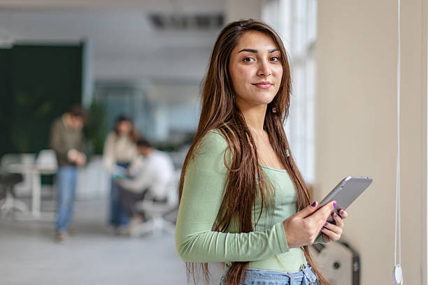 istock Young adult stands in office holding tablet while colleagues work in background on modern tasks during business hours 2252609593