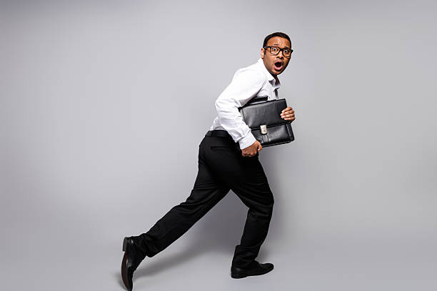 A young handsome businessman in formalwear hurries with a briefcase against a grey background, expressing surprise and urgency. The image conveys work pressure, fast pace, and corporate office dynamics.