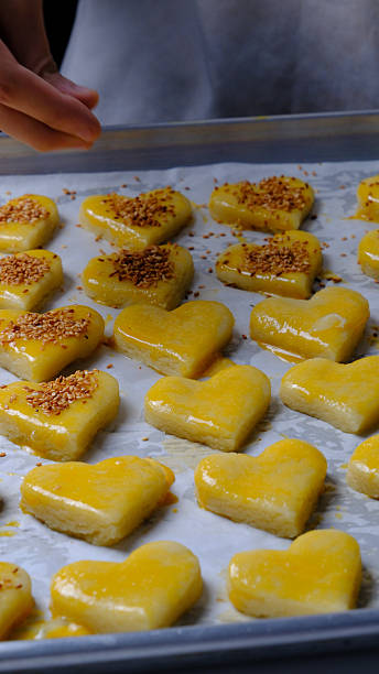 Close-up of a baker’s hand using a silicone brush to apply egg wash onto raw heart-shaped cookie dough arranged on a baking tray before baking.