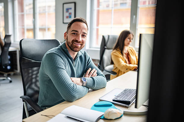 istock Smiling man at desk in modern office while colleague focuses on work in background during business hours 2252626209