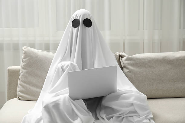 istock Creepy ghost. Person covered with white sheet using laptop on sofa at home 2252629293
