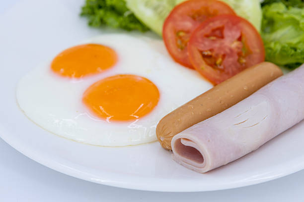 Overhead view of fried eggs served with ham, sausage, fresh salad, tomato, and cucumber on a white plate against white background.