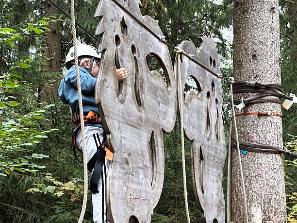 A young boy in a safety helmet and harness carefully climbs across large wooden cut-out panels in an adventure park. Marketing outdoor youth centers, promoting physical activity for kids, or as social media content for travel blogs focusing on family adventures.