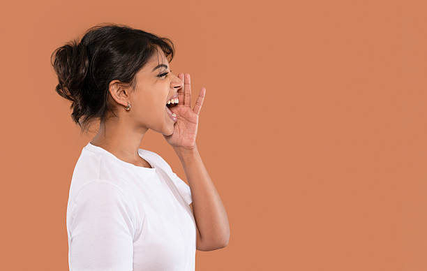 A woman enthusiastically calls out, displaying excitement. She stands against a warm, solid-colored background, showcasing her expressive nature and lively spirit.
