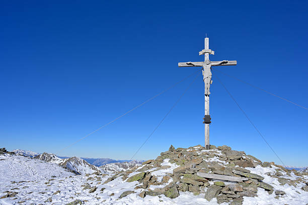 Crucifix on a rocky, snow-covered mountain peak under a clear blue sky. Styria, Austria