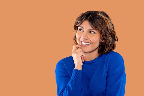 istock Woman in blue sweater smiling while deep in thought against a warm, peach background, radiating positive energy and creativity while engaging in contemplation 2252656529