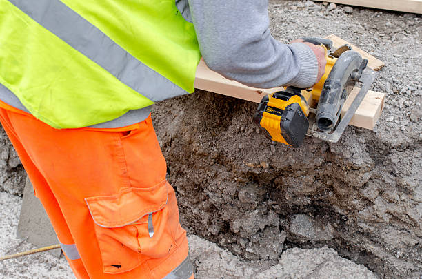 A construction worker in high-visibility gear operates a circular saw to cut through wooden planks on a construction site, surrounded by gravel and dirt.