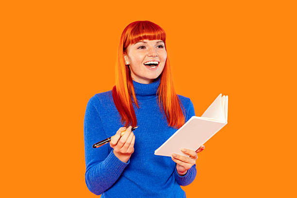 istock Red-haired woman smiling while holding a notebook and pen in a vibrant orange background capturing a moment of inspiration and creativity during a brainstorming session 2252657470