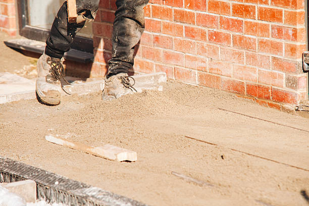 A construction worker is spreading sand to create a leveled base for a patio outside a brick building while wearing safety boots and working in bright daylight.