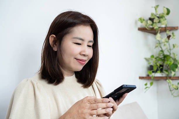 istock A smiling middle-aged Asian woman uses a smartphone while sitting on a sofa in a bright living room. She is texting and browsing apps, 2252660429