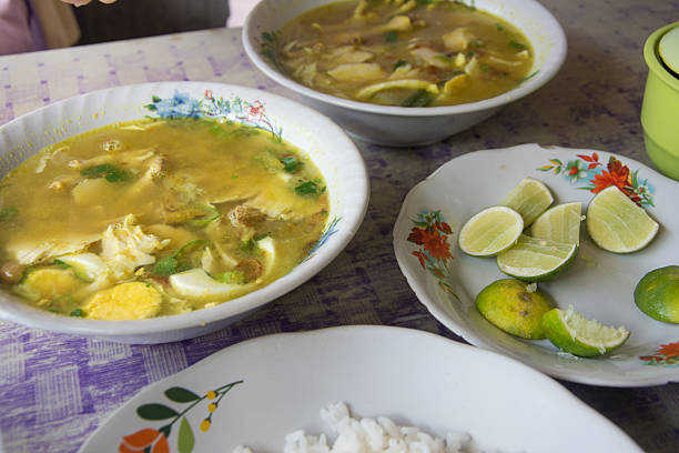 Delicious Indonesian soto ayam chicken soup served in a floral bowl with white rice and fresh lime wedges on a patterned table. Authentic Southeast Asian street food and culinary culture