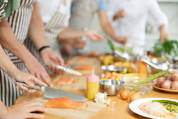 People in aprons standing at table with groceries and utensils during group cooking lesson, absorbed in cooking process, cutting fresh salmon following chef instructions. Cropped shot