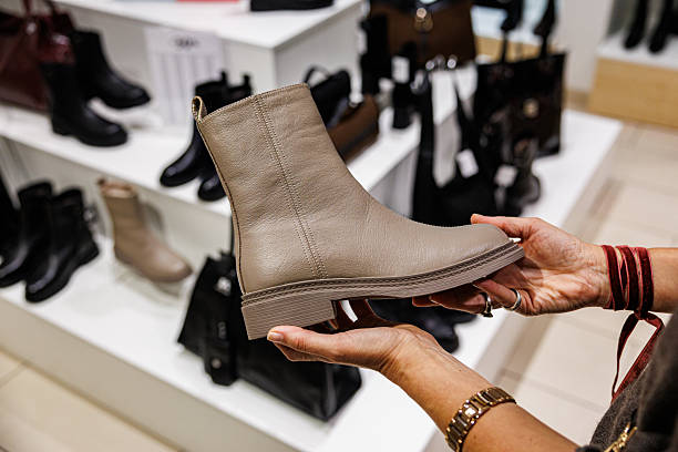 istock A shopper holds a beige leather ankle boot in a retail store, surrounded by various black and brown boots on display. Close-up shot with indoor lighting 2252668580