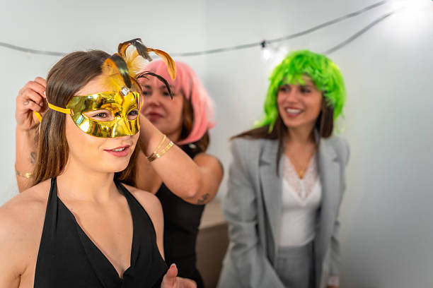 Three young adult women are getting ready for a costume party, with one putting on a golden masquerade mask while her friends watch and smile, wearing colorful wigs in a festive setting