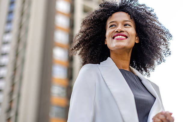 istock Young black woman smiles outdoors in urban setting while wearing a light jacket and standing against tall buildings 2252678545