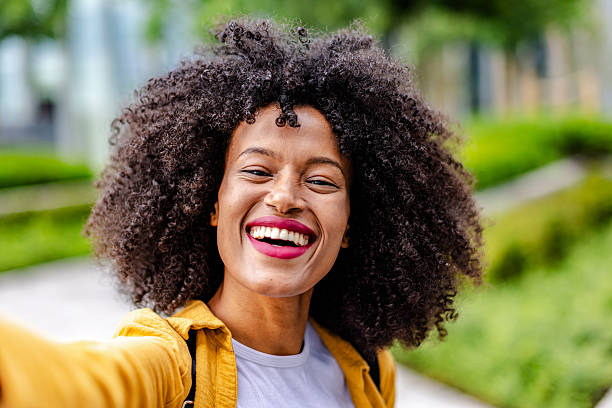 A young black woman stands outside smiling happily while taking a selfie. She enjoys a sunny day surrounded by greenery in a park. Her joy is clear as she captures the moment.
