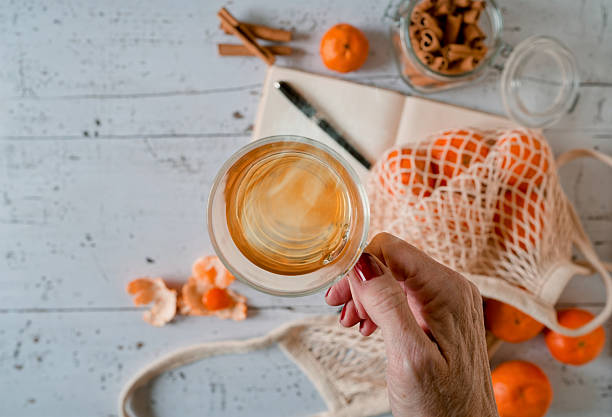 Overhead winter lifestyle flat lay featuring a hand holding a cup of hot tea above a white wooden table with tangerines, cinnamon sticks in a glass jar, an open diary, and a reusable mesh bag. Warm tones and natural textures create a calm seasonal atmosphere with ample copy space for wellness, lifestyle, or winter marketing concepts.
