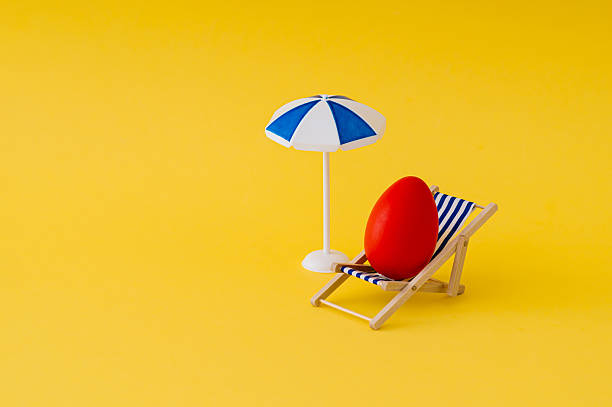 istock Red Easter egg on deck chair with beach umbrella on yellow background. Easter concept. Creative festive holiday layout. Easter aesthetic. Copy space. 2252683628