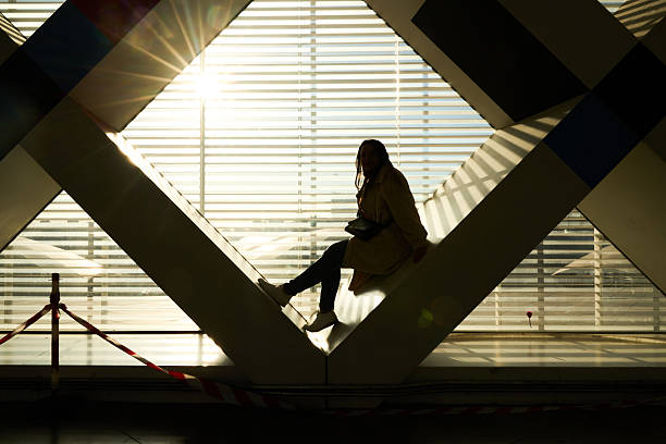 Woman sitting in silhouette against a bright window with sunlight creating geometric shadows on the modern architectural structure