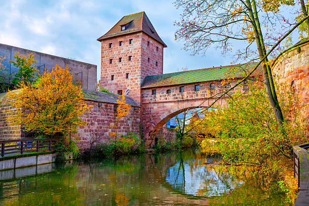 Fronveste building and part of old city wall and bridge system in Nuremberg, Germany, reflected in Pegnitz River. Autumnal scene with historic red sandstone architecture and lush surrounding foliage