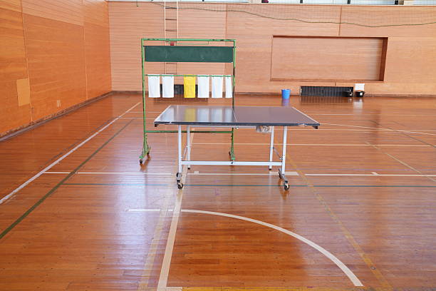 A ping pong table is in a gymnasium with a green board behind it. The table is empty and the gym is empty