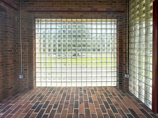 Interior view of a narrow brick hallway featuring a large glass block wall that lets in soft natural light, with red brick floor and side walls creating a geometric, architectural space and a framed view of greenery outside, symbolizing privacy, security and modern design.Ideal for use in architecture and interior design portfolios, renovation and construction marketing, real estate brochures, educational materials about glass block walls, energy efficient daylighting solutions and urban building details.​