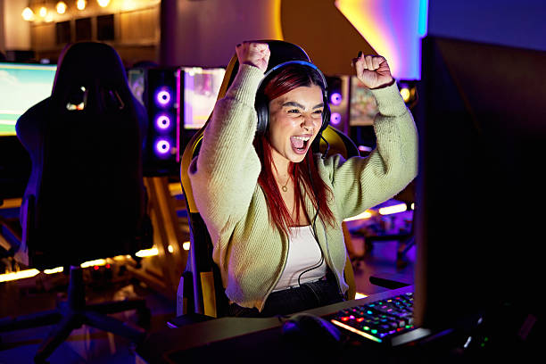 Waist up view of casually dressed woman sitting in gaming chair, wearing headset, yelling and cheering, face illuminated by screen.