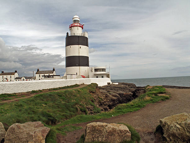 A scenic view of Hook Head Lighthouse standing along the rugged coastline of Ireland. The historic black-and-white lighthouse rises above rocky shores, overlooking the surrounding sea. This image highlights Ireland’s maritime heritage and the dramatic coastal landscapes that have guided sailors for centuries.