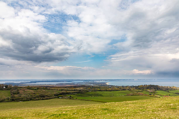 Swanage. Ballard Cliff path, Dorset. A peaceful coastal landscape with green fields and rolling hills fading into a distant sea beneath a dramatic, cloud-filled sky.