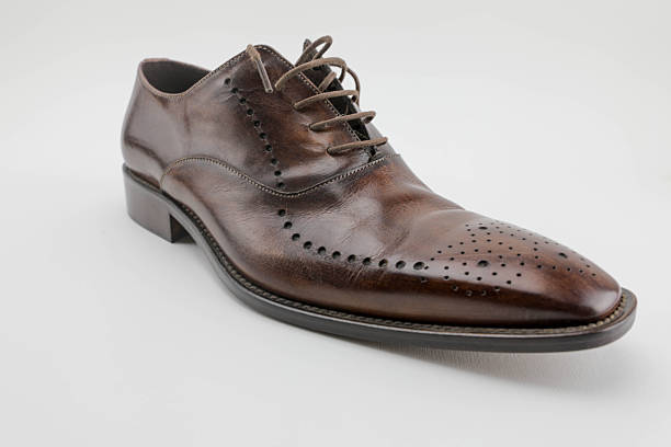 Single Brown Leather Oxford Shoe With Elegant Brogue Detailing, Photographed On A Clean White Background. The Polished Surface, Classic Stitching, And Refined Shape Emphasize Sophisticated Men’s Fashion, Formal Footwear, And Timeless Craftsmanship.