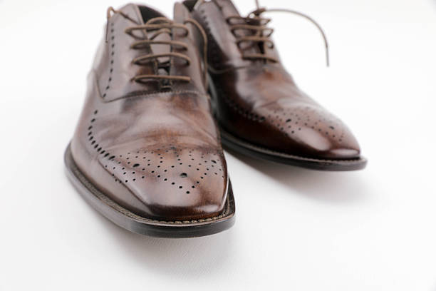 Close-Up Of Elegant Brown Leather Oxford Shoes With Decorative Brogue Detailing, Placed On A Clean White Background. The Polished Surface And Classic Design Highlight Timeless Men’s Fashion, Formal Wear, And Traditional Craftsmanship.