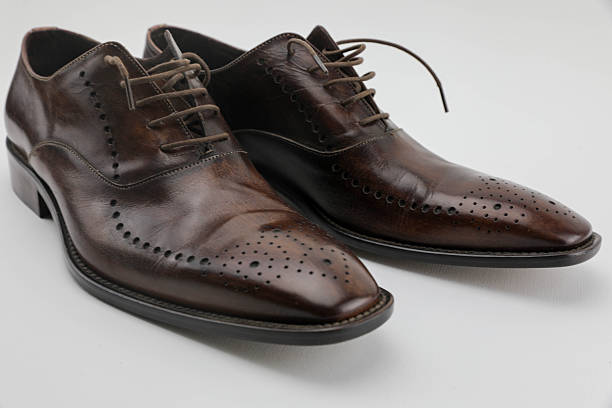 Close-Up Of Elegant Brown Leather Oxford Shoes With Decorative Brogue Detailing, Placed On A Clean White Background. The Polished Surface And Classic Design Highlight Timeless Men’s Fashion, Formal Wear, And Traditional Craftsmanship.