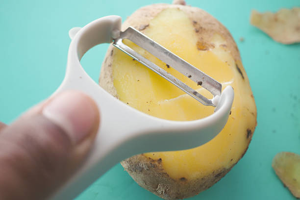 A multiracial woman wearing a yellow sweater and jeans peels potatoes on a cutting board in a bright kitchen while preparing baby food.