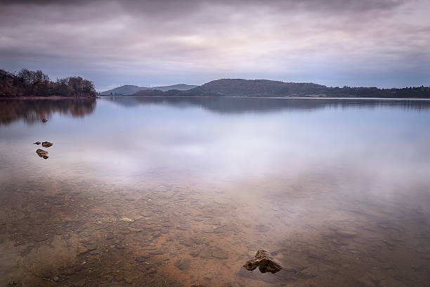 Calm water at Urrunaga reservoir in Legutio with rocks in foreground and misty mountains at dusk