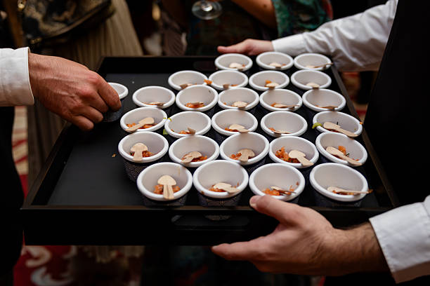 Waiter's hands in uniform hold a tray of elegant mini cups and wooden spoons with bite-size appetizers being served to guests at a formal event or reception