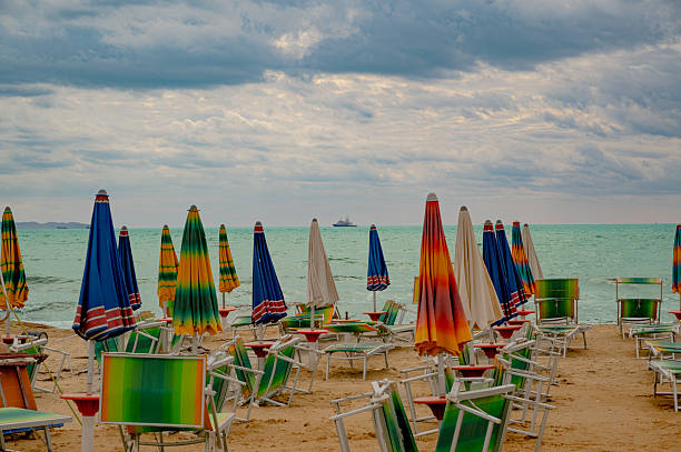 Colorful folded beach umbrellas placed near sun loungers on an empty sandy beach by the cold winter sea in Albania, conveying seasonal contrast, calm, and off-season coastal atmosphere.