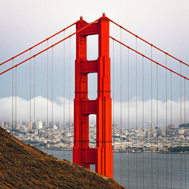 Close-up view of a bright red tower of the Golden Gate Bridge rising above San Francisco Bay, with the city skyline visible in the background under soft clouds. This iconic suspension bridge represents American engineering, travel, and urban identity, making it ideal for themes of architecture, transportation, tourism, and famous landmarks.