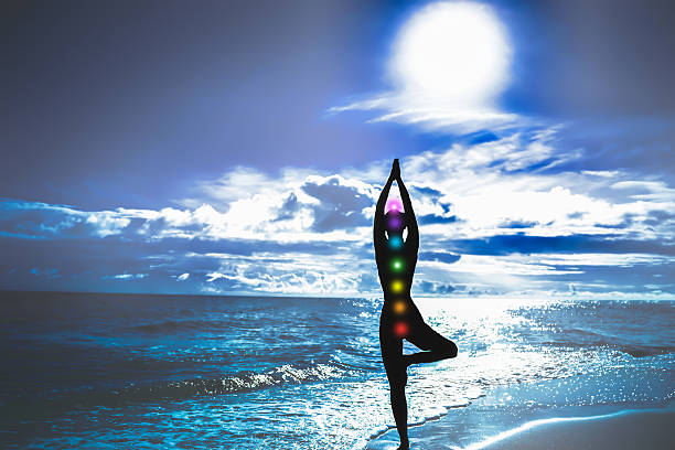 istock Woman silhouette with chakras practicing yoga tree pose on a serene blue beach at night. 2252754161